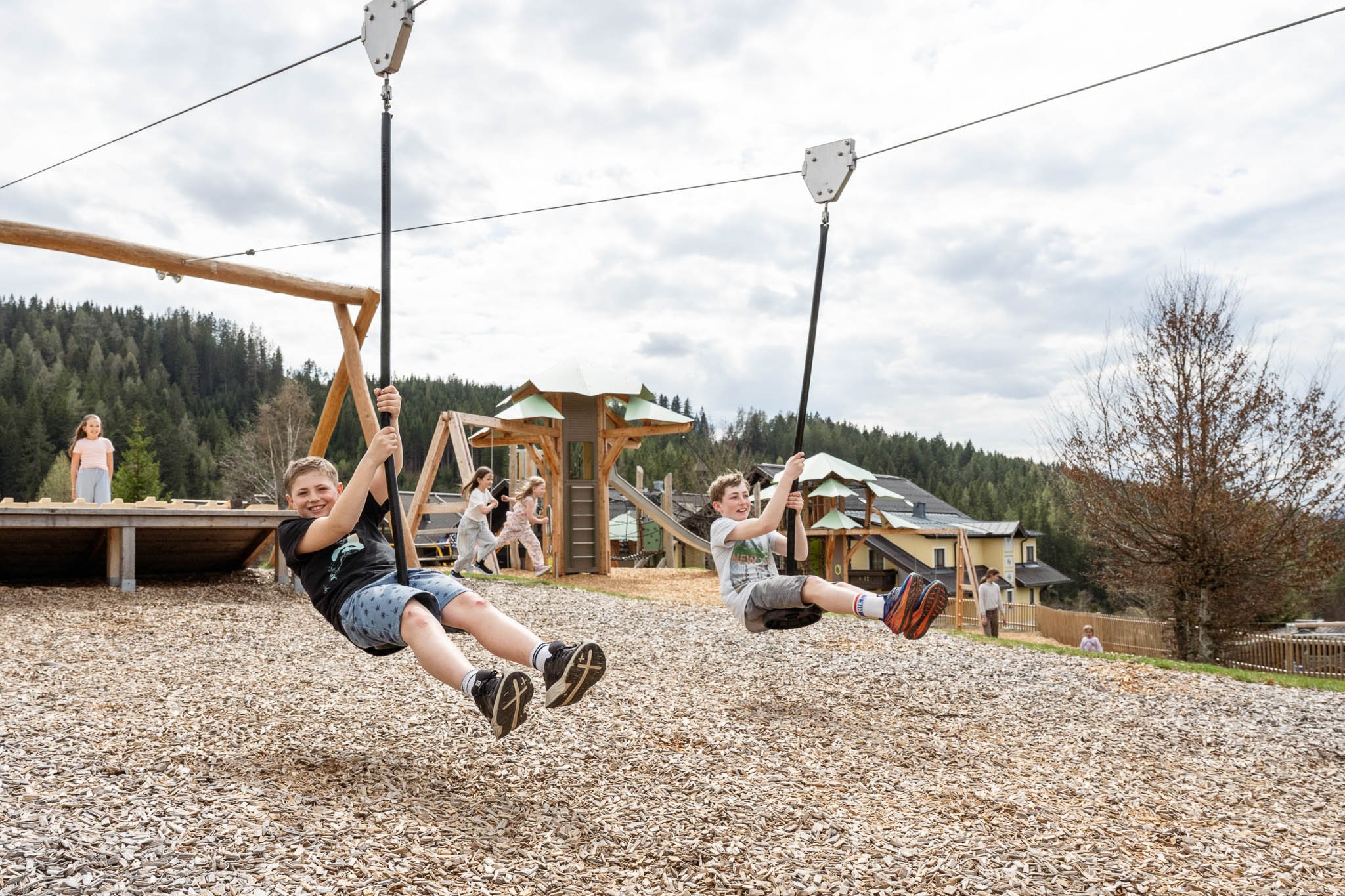 Zwei Jungen fahren auf einer Seilrutsche auf einem Spielplatz, im Hintergrund spielen Kinder und Bäume
