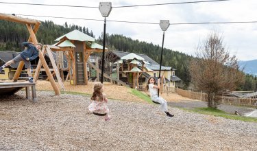 Kinder spielen auf einer Seilrutsche und Klettergeräten auf einem Spielplatz vor Wald und Häusern