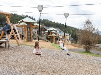 Kinder spielen auf einer Seilrutsche und Klettergeräten auf einem Spielplatz vor Wald und Häusern