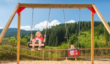 Zwei Kinder schaukeln auf einem Spielplatz, umgeben von Bäumen und einem Zaun.