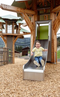 Kind rutscht lachend auf einer Metallrutsche eines Holzspielplatzes mit Baumhaus im Hintergrund
