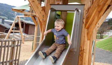 Kleiner Junge rutscht lachend eine Metallrutsche auf einem Holzspielplatz hinunter
