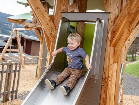 Kleiner Junge rutscht lachend eine Metallrutsche auf einem Holzspielplatz hinunter