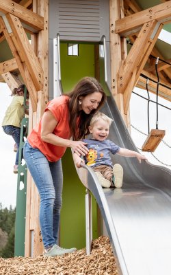 Frau hilft lachendem Kind auf einer Rutsche aus Metall auf einem Holzspielplatz