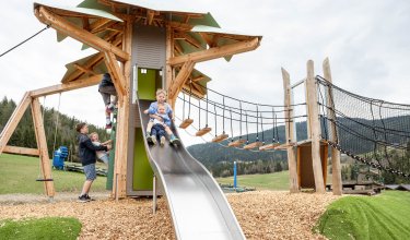 Kinder rutschen und klettern an einem großen Holzspielplatz mit Rutsche und Hängebrücke im Freien