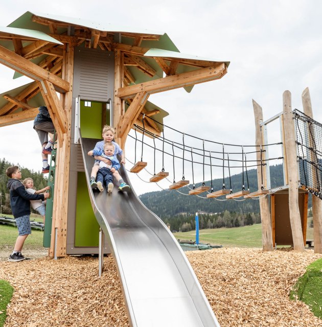 Kinder rutschen und klettern an einem großen Holzspielplatz mit Rutsche und Hängebrücke im Freien