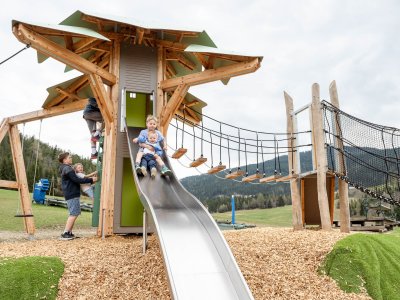 Children sliding and climbing on a large wooden playground with slide and rope bridge outdoors