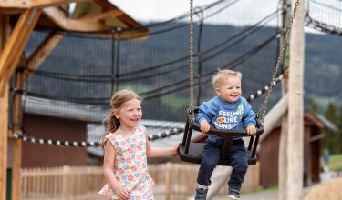 Mädchen schiebt lachendes Kleinkind auf Schaukel auf einem Spielplatz mit Holzkonstruktion
