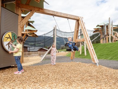 Kinder spielen auf einem Holzspielplatz mit Schaukel und Kletternetz in ländlicher Umgebung