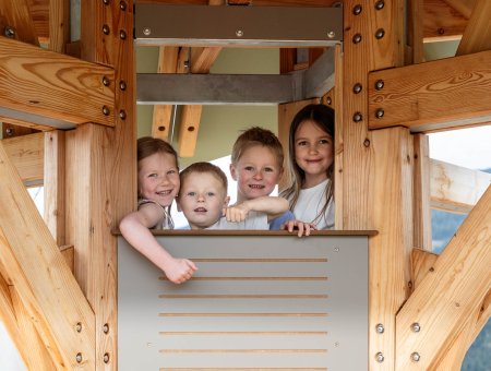 Vier Kinder schauen aus einem Holzspielhausfenster und lächeln in die Kamera