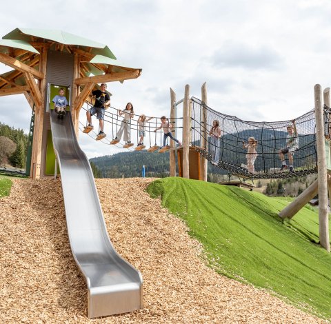 Children playing on a wooden climbing structure with slide and net bridge in a mountain setting