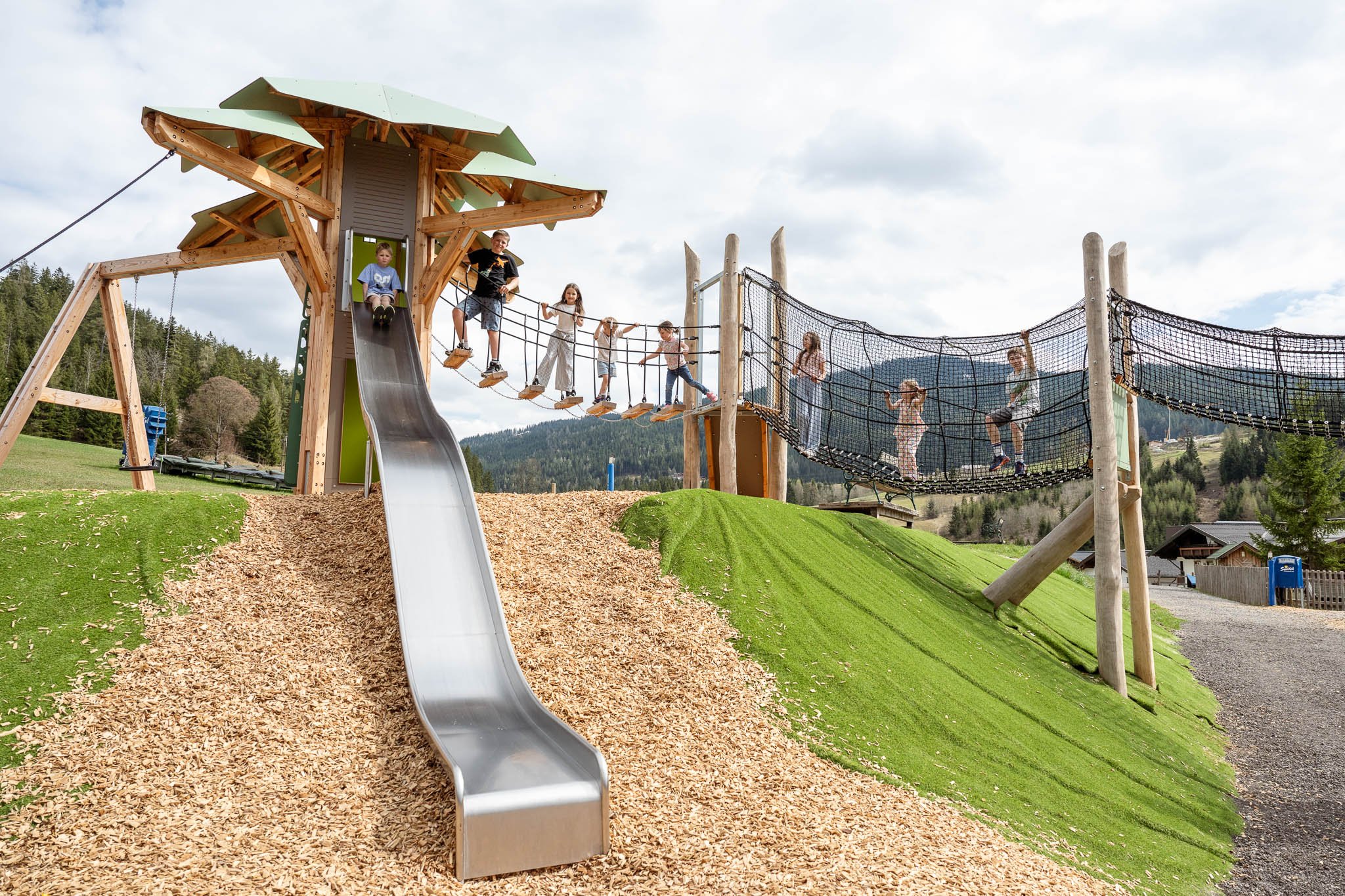 Kinder spielen auf einem Klettergerüst mit Rutsche und Netzbrücke in einer Berglandschaft