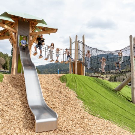 Kinder spielen auf einem Klettergerüst mit Rutsche und Netzbrücke in einer Berglandschaft