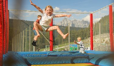 Kinder springen fröhlich auf einem Trampolin mit Netz und Bergen im Hintergrund
