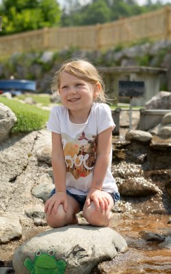 Mädchen kniet lächelnd an einem Bach mit Spielfrosch