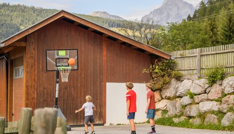 Drei Jungen spielen Basketball vor einer Holzhütte mit Bergblick im Hintergrund.