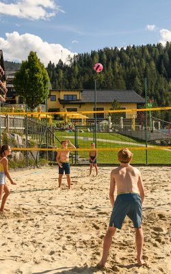 Kinder spielen Beachvolleyball auf Sandplatz mit gelbem Netz vor Wald und Häusern
