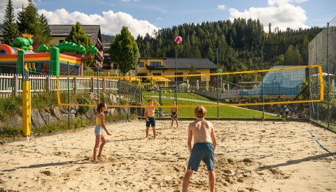 Kinder spielen Beachvolleyball auf Sandplatz mit gelbem Netz vor Wald und Häusern