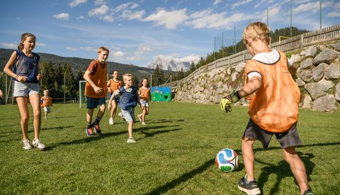 Kinder spielen Fußball auf einem grünen Feld vor einer Steinmauer und Bergen im Hintergrund