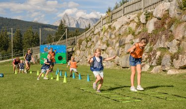 Kinder spielen draußen auf einer Wiese mit Hütchen und Kletterleiter vor einem Felsen und Bergen im Hintergrund