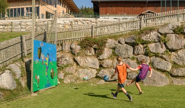 Zwei Jungen spielen Fußball auf einer Wiese vor einer bemalten Torwand mit Löchern