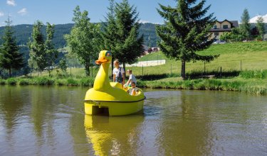 Kinder in einem gelben Enten-Pedalboot auf einem See.