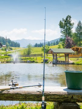 Angelrute und Eimer auf einer Bank mit See und Hütte im Hintergrund.