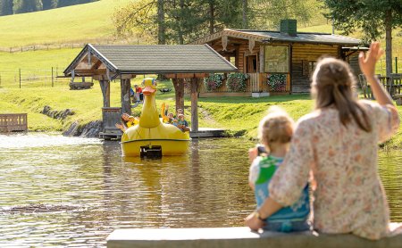 Mutter und Kind winken einem Tretboot in Entenform auf dem Teich zu.