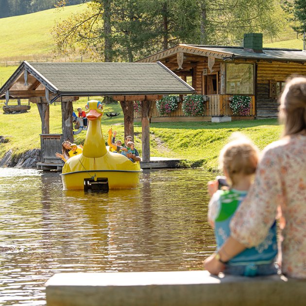 Mutter und Kind winken einem Tretboot in Entenform auf dem Teich zu.