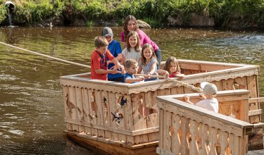 Kinder ziehen ein Holzfloß mit einem Seil über das Wasser.