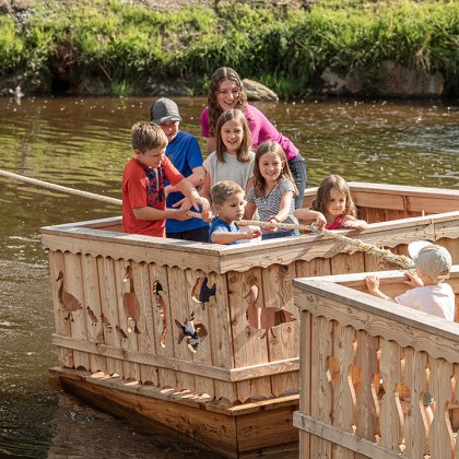 Kinder ziehen ein Holzfloß mit einem Seil über das Wasser.
