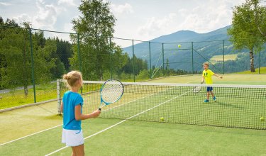 Zwei Kinder spielen Tennis auf einem Außenplatz mit Bergblick.