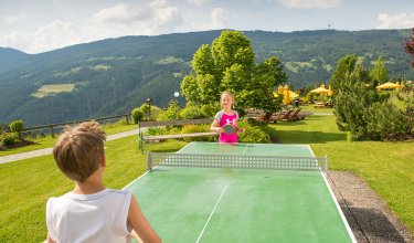 Zwei Kinder spielen Tischtennis im Freien mit Bergkulisse.