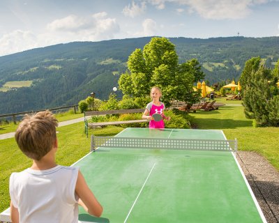 Zwei Kinder spielen Tischtennis im Freien mit Bergkulisse.