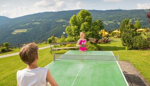Zwei Kinder spielen Tischtennis im Freien mit Bergkulisse.