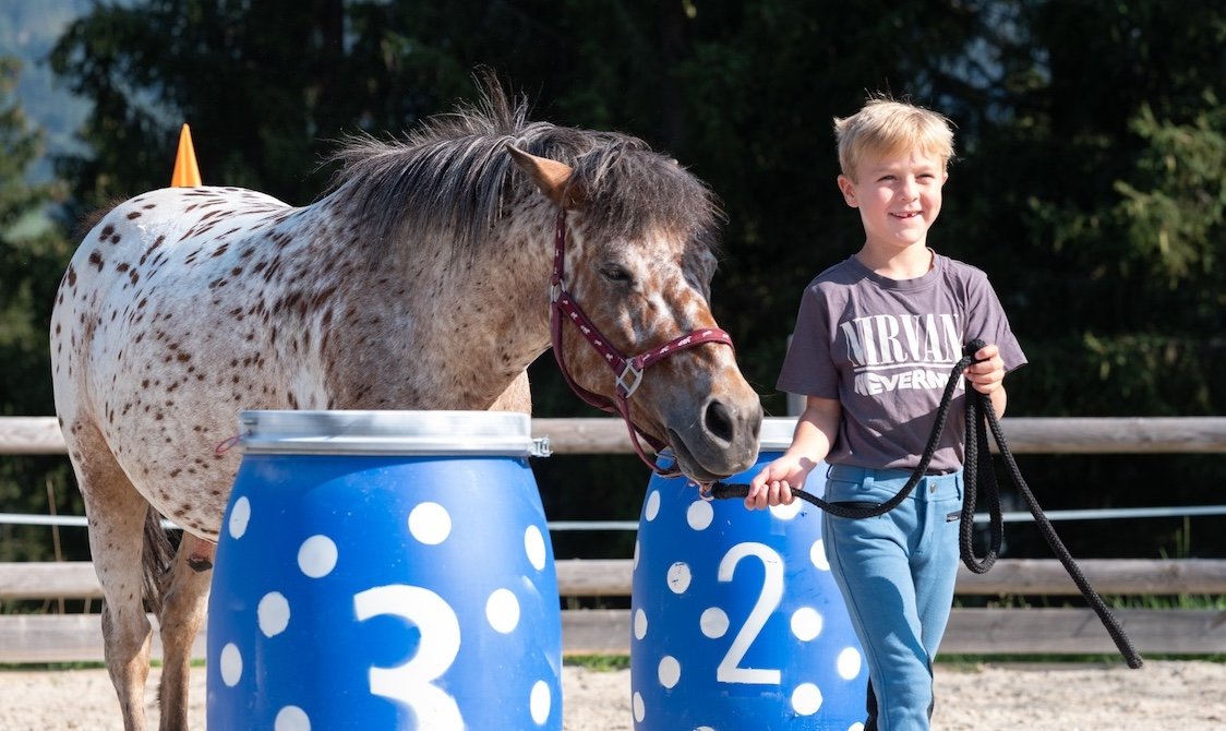 Junge führt Pony an der Leine neben blauen Fässern mit den Zahlen 2 und 3