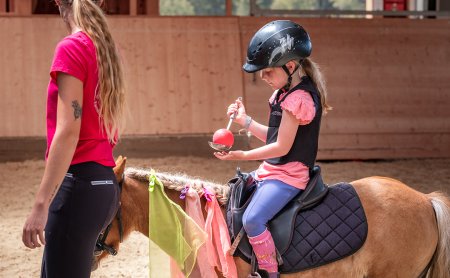 Kind mit Helm sitzt auf Pony und hält einen Löffel mit rotem Ball, daneben steht eine Frau