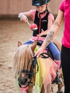 Kind mit Helm auf Pony sitzt, hält roten Ball mit Schöpfkelle, neben Person mit Tattoo und pinkem Shirt