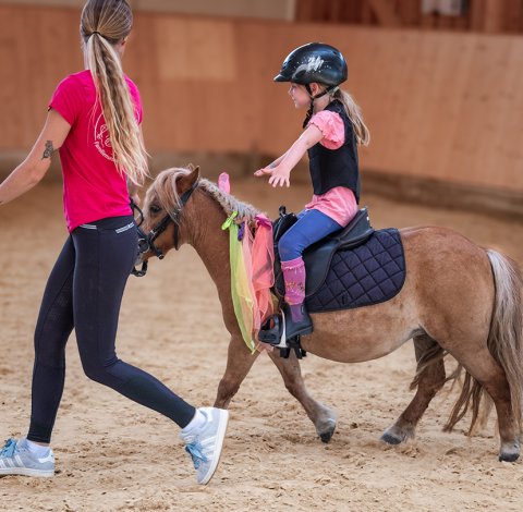 Kind mit Helm und Schutzweste reitet Pony in Reithalle, Frau führt Pony an der Hand