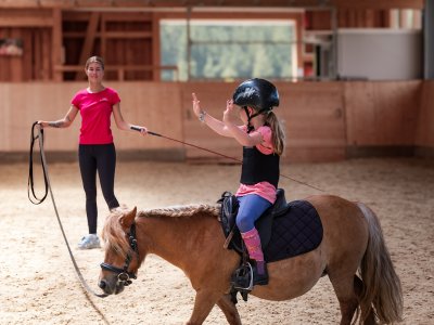 Kind mit Helm und Schutzweste sitzt auf Pony, Trainerin hält Führstrick in Reithalle