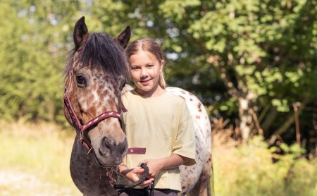 Mädchen steht neben einem gescheckten Pferd im Freien vor grünen Bäumen