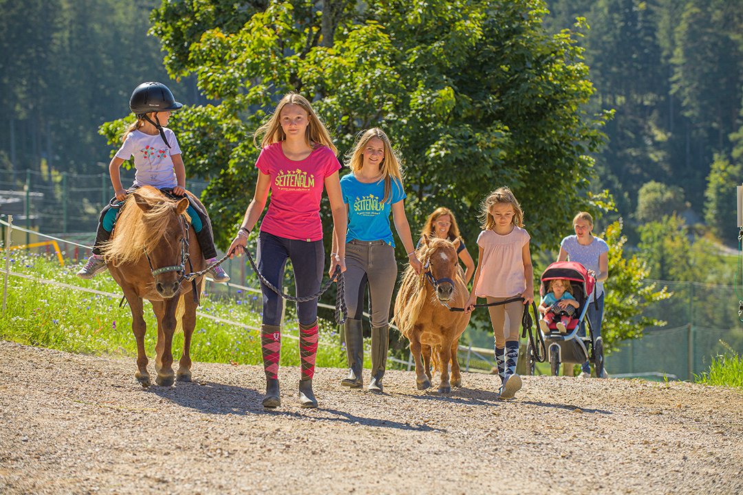 Kinder reiten und führen Ponys auf einem sonnigen Schotterweg.
