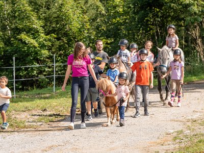 Kinder und Erwachsene führen gemeinsam Ponys auf einem sonnigen Waldweg.
