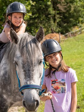 Ein Mädchen reitet ein graues Pony, das andere führt es lächelnd am Strick.