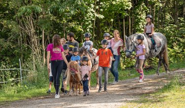 Kinder und Betreuer führen Ponys auf einem Waldweg beim Gruppenausritt.