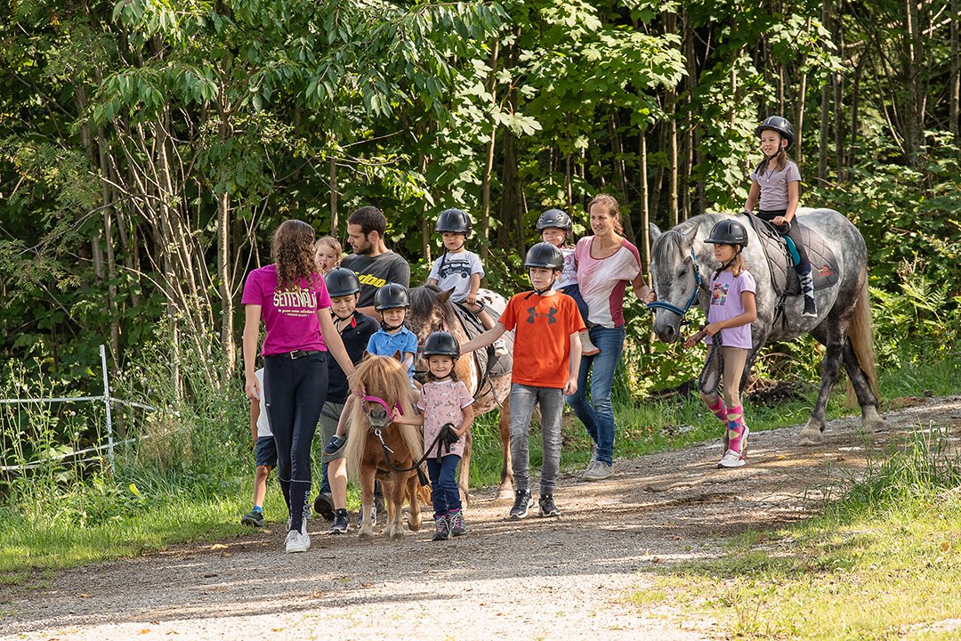 Kinder und Betreuer führen Ponys auf einem Waldweg beim Gruppenausritt.
