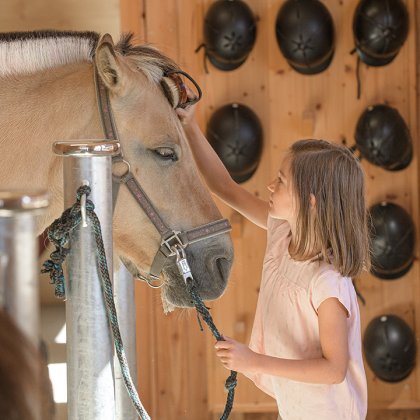 Mädchen streichelt Pony am Anbindeplatz mit Reithelmen im Hintergrund.