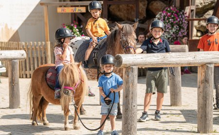 Kinder reiten Ponys und führen sie im sonnigen Hof.