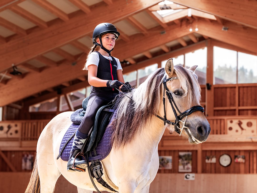 Mädchen mit Helm reitet Pferd in einer hellen Reithalle mit Holzbalken