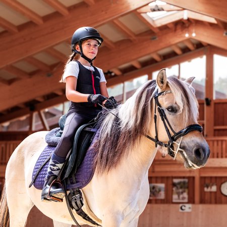 Mädchen mit Helm reitet Pferd in einer hellen Reithalle mit Holzbalken